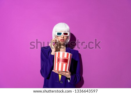 Amazed woman in wig and 3d glasses eating popcorn on purple background