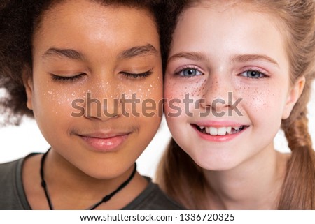 Standing closely. Appealing flawless kids connecting cheeks and displaying their faces with fake freckles