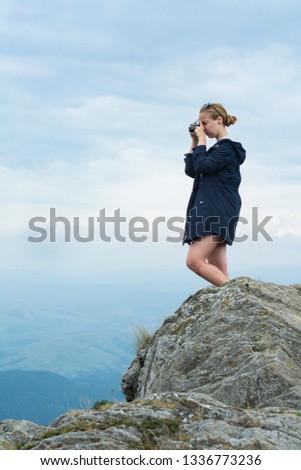 A girl tourist stands on a rock over a cliff in the Carpathians and photographs the landscape.