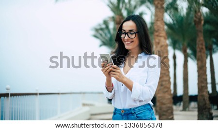 Sending a little of joy. Girl in a calm mood is using her smartphone for talking to someone while standing near the quay among the vibrant green palms and evening clear blue sky.