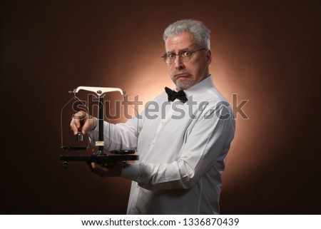 Elderly gray-haired man 50s, in white shirt, glasses and bow tie weighing something on scales with small kettlebells