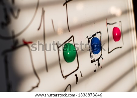 magnets in green, blue, red on the board marker, in the squares drawn by the maker. the light from the window falls on the board leaving a shadow from the blinds. sharpness on the second magnet blue