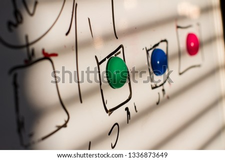 magnets in green, blue, red on the board marker, in the squares drawn by the maker. the light from the window falls on the board leaving a shadow from the blinds. sharpness on the first magnet green 