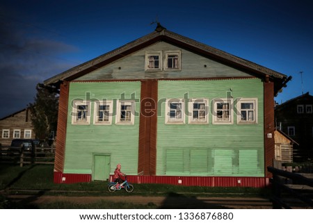 Renovated greena and brown wooden house in rural Russia with a girl on bike riding in front and anthlers placed on top of the building.