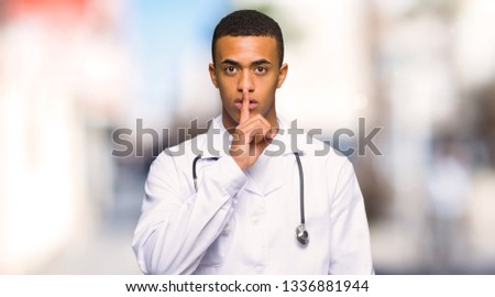 Young afro american man doctor showing a sign of silence gesture putting finger in mouth at outdoors