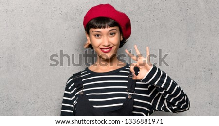 Young woman with beret showing ok sign with fingers over textured wall