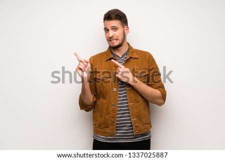 Handsome man over white wall frightened and pointing to the side