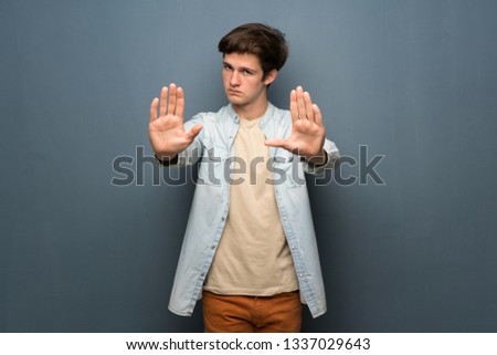 Teenager man with jean jacket over grey wall making stop gesture and disappointed