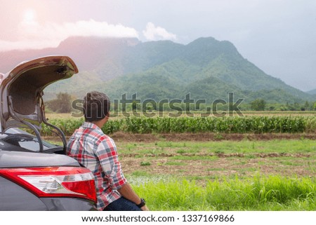 Young man sits in the car trunk looking nature  and relax with green corn field and mountain nature background. Summer vocation with car on the road concept.