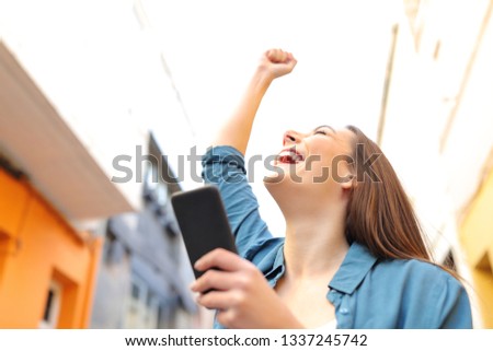 Excited woman holding smart phone celebrates success in the street