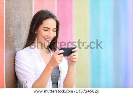 Happy girl watching videos on smart phone leaning on a wall in a colorful street