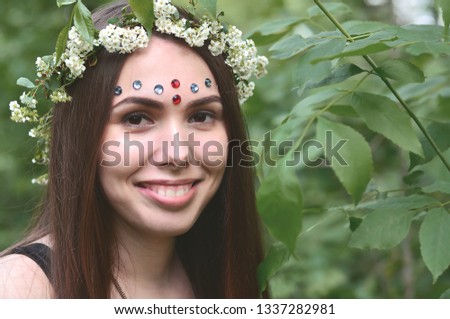 A forest picture of a beautiful young brunette of European appearance with dark brown eyes and large lips. On the girl's head is wearing a floral wreath, on her forehead shiny decorations