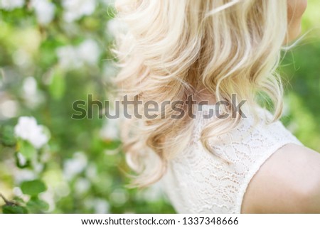 Fashion outdoor photo of beautiful young woman  posing in garden with blossom trees. Blonde in flowering gardens. Beautiful streaming blonde curls.  View from a back. Close up picture
