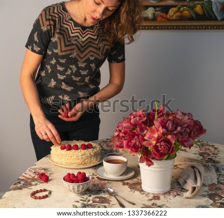 Moscow/Russia-CIRCA 03.2019: an image of a female decorating a homemade cake with raspberries, cup of tea and bouquet of red color blossomed tulips on a table