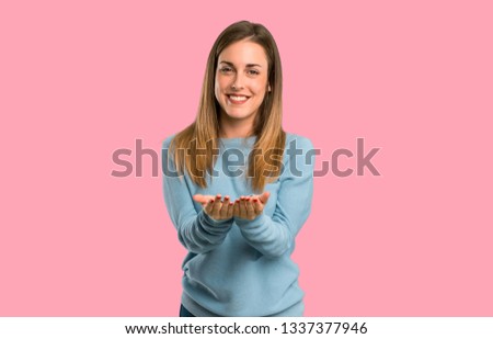 Blonde woman with blue shirt holding copyspace imaginary on the palm to insert an ad on isolated pink background