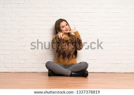 Teenager girl sitting on the floor making phone gesture and pointing front