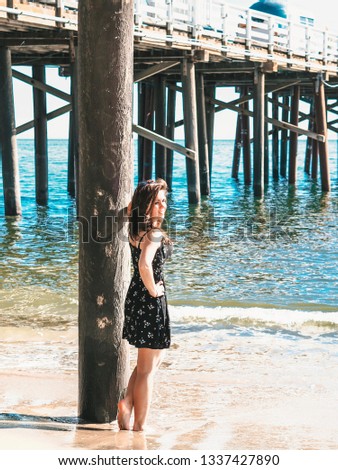 Brunette girl in short dress stands leaning on a beam on Malibu pier in Los Angeles