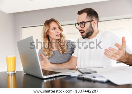 Image of displeased young loving couple sitting at the kitchen using laptop computer.
