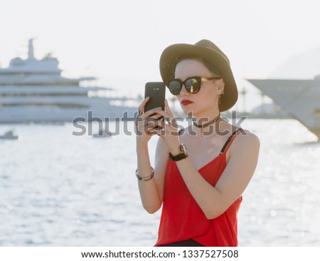 Girl in sunglasses and a black hat on the background of yachts in the resort European city takes pictures on a smartphone or takes a selfie. Travel and Vacation