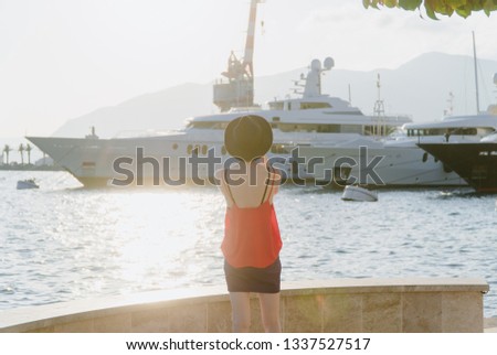 A girl in sunglasses and a black hat is standing against the background of yachts in a European resort town looking into the distance. Shot from behind. Travel and Vacation