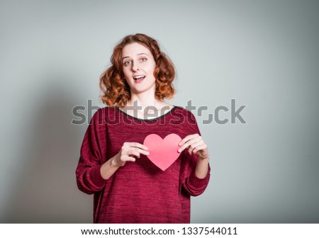 Cute redhead girl holds paper heart - symbol of love, isolated on gray background