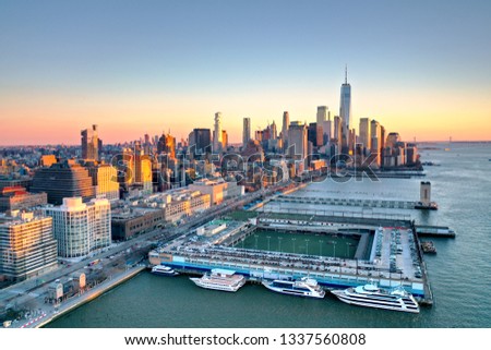Photo shows New York Manhattan skyline at with vanilla twilight sky during the sunset hour. The photo also show the high rise buildings with a football pitch and some cruise boats parked at the pier.