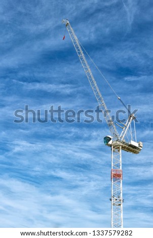 A White Tower Crane with in the Blue Skies with light layered clouds looking isolated with the sunlight illuminating the Structure. Copy Space to the left of the image.
