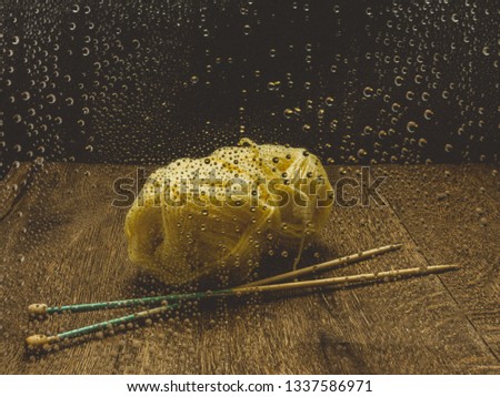 Yellow wool and knitting needles on a wooden surface, seen through a window with rain drops. 