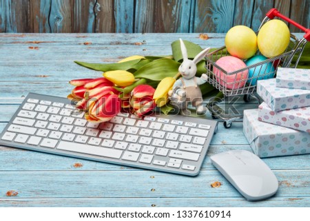 Colored easter eggs in shopping trolley, gift boxes,  tulips flowers  and  computer keyboard and mouse on a wooden  background. Top view. Easter background, Easter shopping.