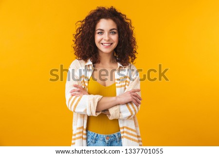 Image of young woman 20s with curly hair smiling and standing with arms crossed, isolated over yellow background