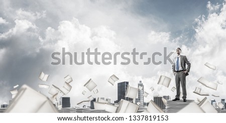 Confident businessman in suit standing on pile of documents among flying books with cloudly sky on background. Mixed media.