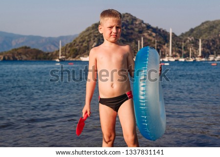 a six year old tanned boy stands holding a swimming circle and a sand shovel by the sea in Turkey