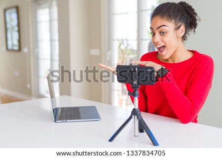 Young african american woman doing video call using smartphone camera amazed and smiling to the camera while presenting with hand and pointing with finger.