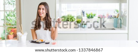 Wide angle picture of beautiful young woman eating asian sushi from delivery with a big smile on face, pointing with hand and finger to the side looking at the camera.