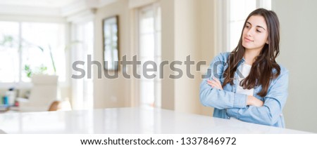 Wide angle picture of beautiful young woman sitting on white table at home smiling looking to the side with arms crossed convinced and confident Royalty-Free Stock Photo #1337846972