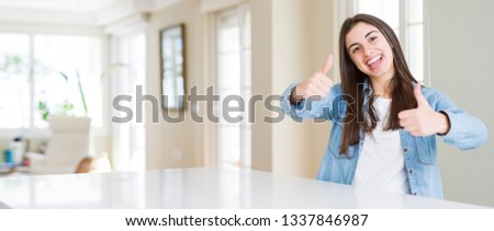Wide angle picture of beautiful young woman sitting on white table at home approving doing positive gesture with hand, thumbs up smiling and happy for success. Looking at the camera, winner gesture.