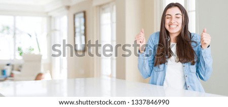 Wide angle picture of beautiful young woman sitting on white table at home excited for success with arms raised celebrating victory smiling. Winner concept.