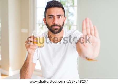 Handsome hispanic man drinking healthy orange juice with open hand doing stop sign with serious and confident expression, defense gesture