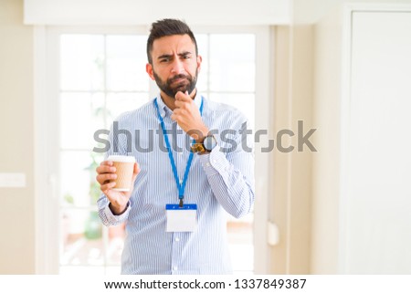Handsome hispanic man wearing id card and drinking a cup of coffee serious face thinking about question, very confused idea