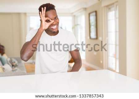 Handsome african american man on white table at home doing ok gesture with hand smiling, eye looking through fingers with happy face.