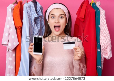 Photo of screaming young lady in clothes store. Girl with opened mouth holds mobile phone and credit card. Female near hagers with colourful outfits. Woman gets message with ballance on card.