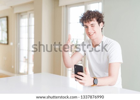 Young man using smartphone at home happy with big smile doing ok sign, thumb up with fingers, excellent sign