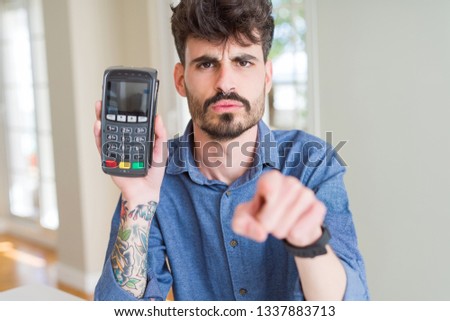 Young man holding dataphone point of sale as payment pointing with finger to the camera and to you, hand sign, positive and confident gesture from the front