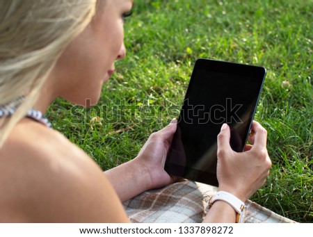 A young girl uses a tablet outdoors lying on green grass.