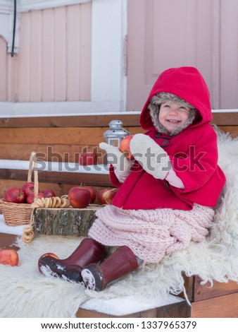 Two years old child in red coat at winter. Russian style village photo. Red apples in basket and snow