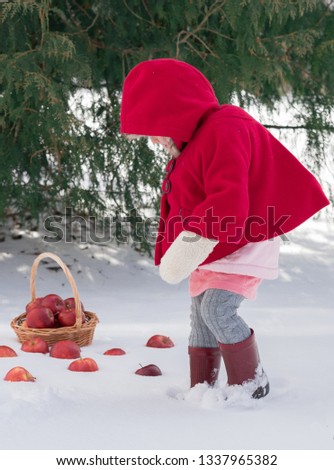 Two years old child in red coat at winter. Russian style village photo. Red apples in basket and snow