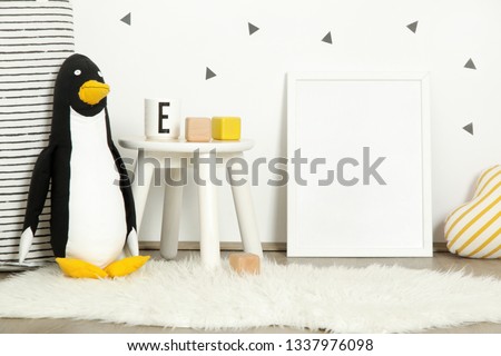 Blank white poster in the cute kid's corner, blank framed poster leaning on the wall next to the wooden chair and kids toys
