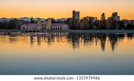 A clear, golden reflection of the City Park boathouse in a sparkling blue pond during a vibrant yellow sunset in the late summer or early autumn. Denver, Colorado, USA.