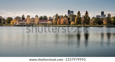 A beautiful panorama of City Park in Denver, Colorado in late summer or autumn. The brick boathouse and a row of trees changing color are reflected in the smooth, blue, peaceful water of the pond.