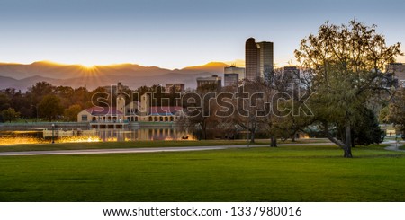 Panorama photo of City Park in Denver, Colorado at sunset. Sun rays shine down on the sparkling golden pond, boathouse and a wide green lawn with office buildings and layers of purple mountains.